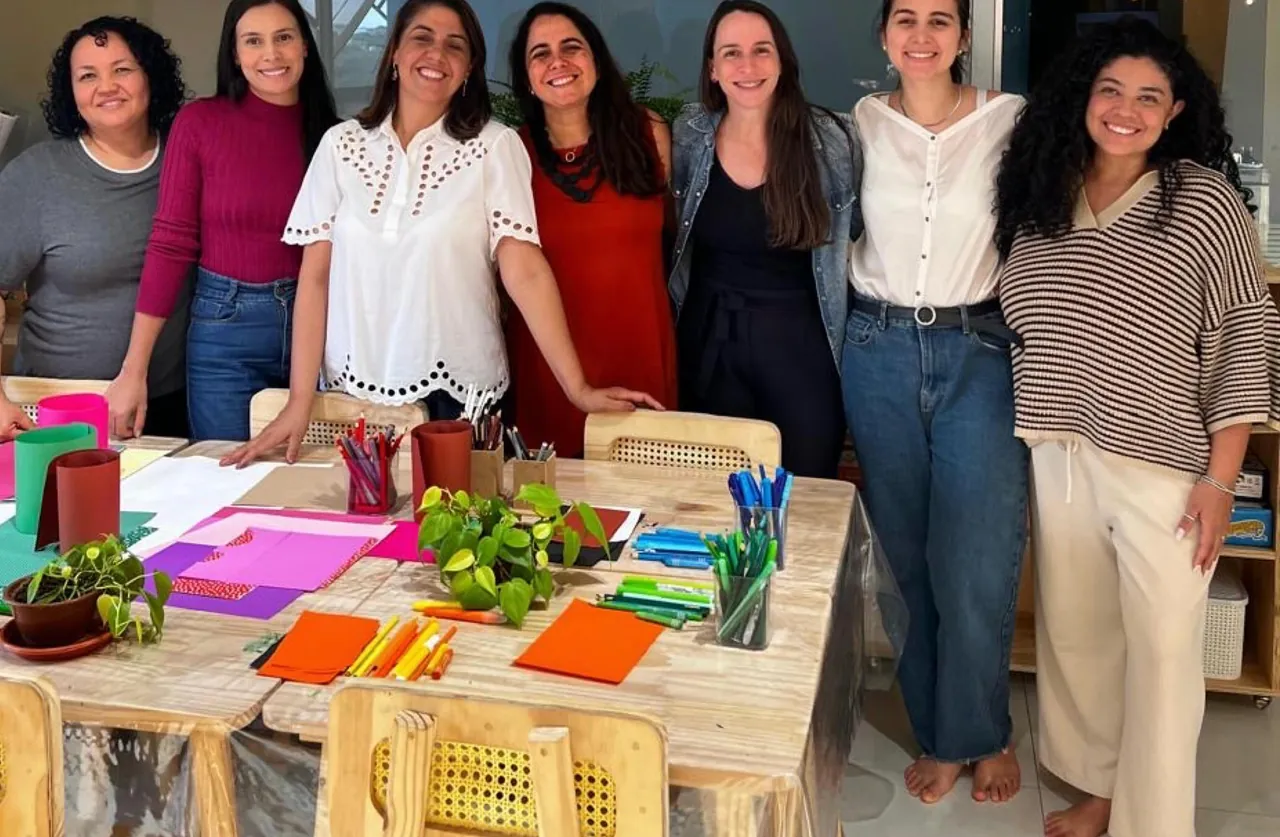 Sete educadoras sorrindo em pé atrás da mesa de projetos, coberta de papéis coloridos, lápis e plantas